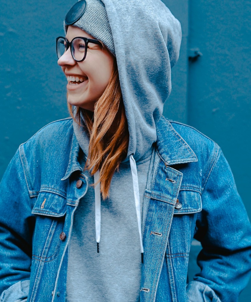 a lady with long hair and glasses, wearing a hoodie and a denim jacket. She is looking off to her right.