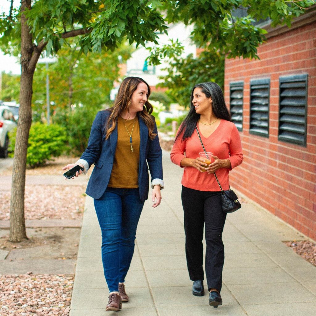 two women walking along a quiet street. They are looking at each other and talking. They are professionally dressed and seem to be in a good mood.