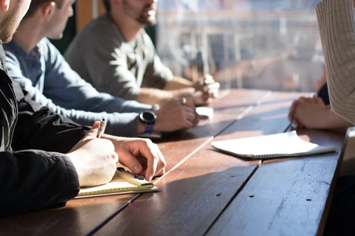 4 people sitting around a table meeting. They are casually dressed and hold notepads and pencils. We can't really see their faces.